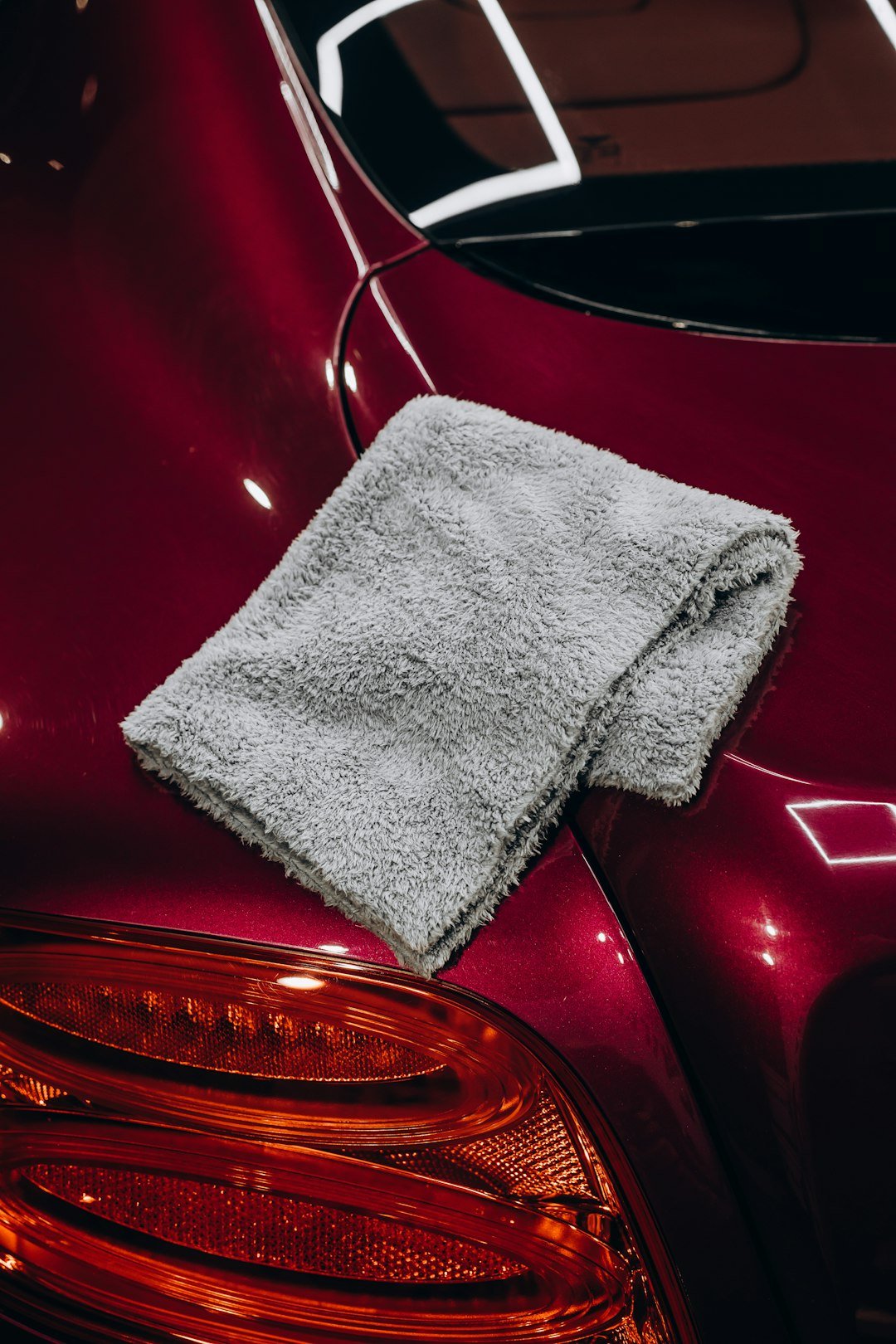 A folded grey plush microfiber towel sits on the gleaming trunk of a modern red car after a professional detail. The flawless, glossy paint reflects the overhead lights, showcasing the results of polishing and paint correction. Image is suitable for themes of automotive care, luxury vehicle maintenance, detailing services, and car cleaning products.